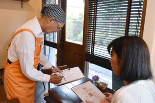 認知症の人が接客するカフェ「オレンジデイ　ＳＥＮＧＡＷＡ」で注文を取る森田利雄さん（左）＝９月１３日、東京都調布市